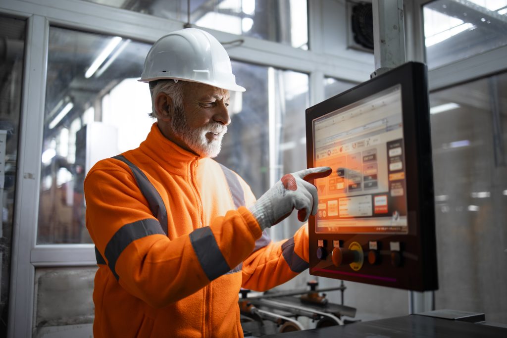 An experienced industrial engineer or senior factory worker operating automated machine in control room of production hall. Factory interior in background.