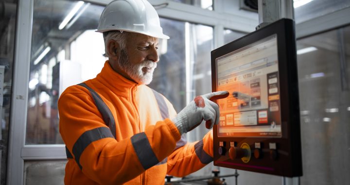 An experienced industrial engineer or senior factory worker operating automated machine in control room of production hall. Factory interior in background.